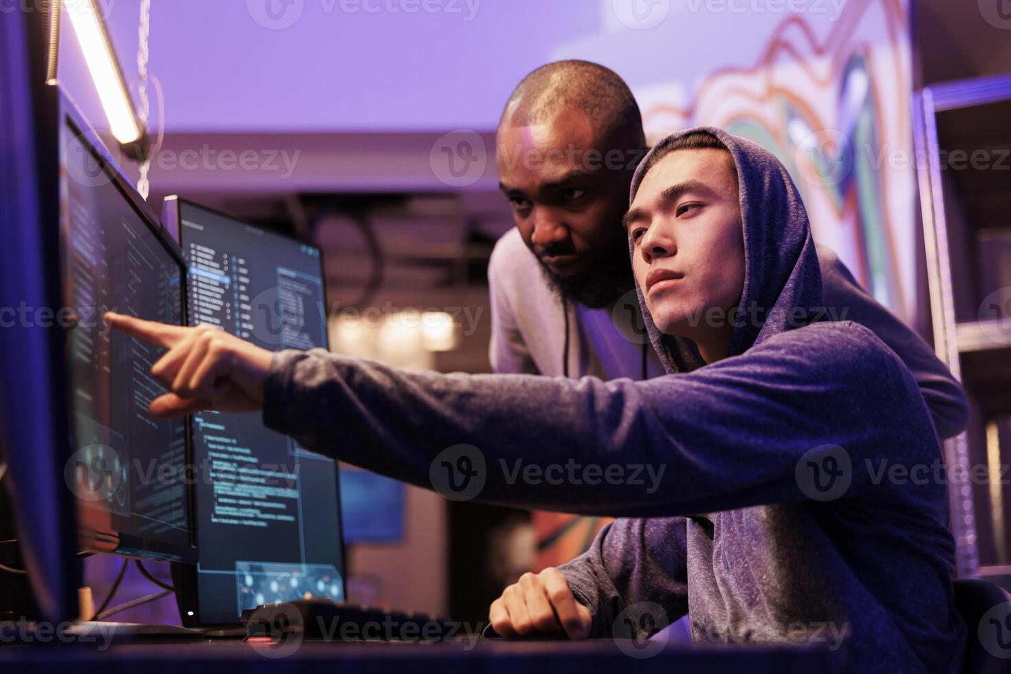 Hacker pointing at malware on screen while working together in abandoned warehouse. Asian criminal in hood showing code lines on computer to ransomware developer while cracking password photo