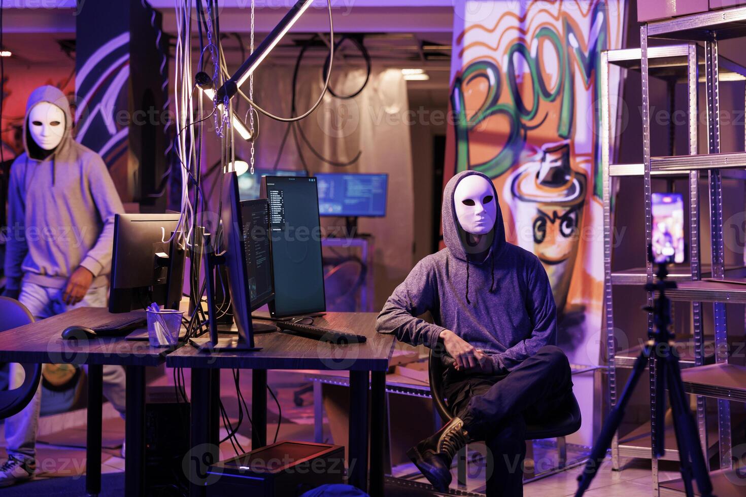 Anonymous hacker in mask recording threat video on smartphone camera in abandoned warehouse. Cybercriminals breaking law while streaming internet fraud and cyberattack on mobile phone photo