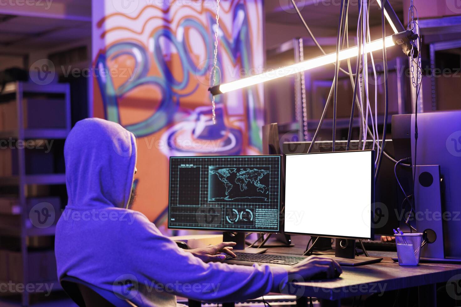 Criminal in hood coding malicious software on computer with empty white screen and hacking system. Internet scammer planning phishing attack while using monitor with blank display photo