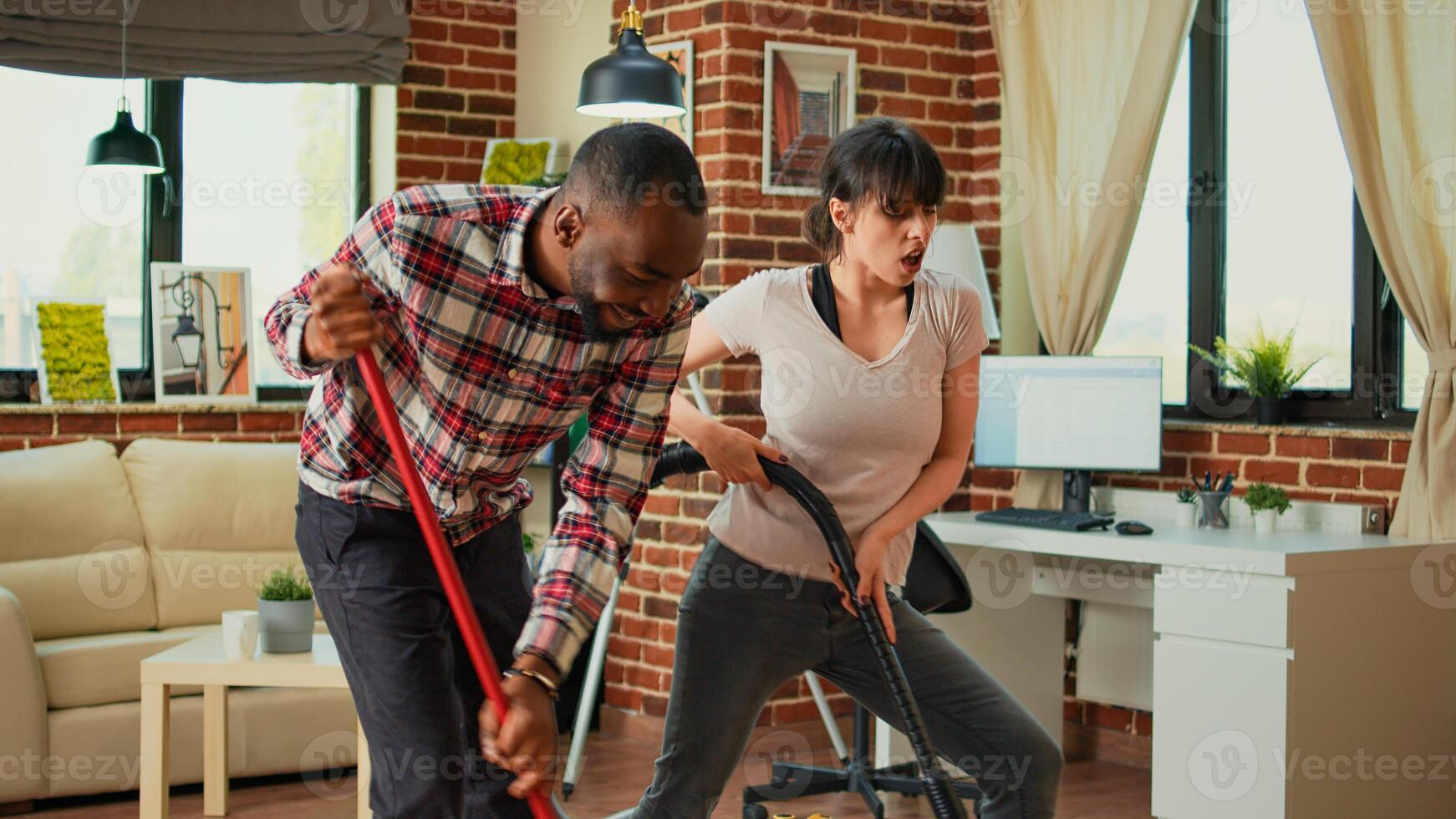 Diverse couple showing dance moves and having fun cleaning apartment, using mop to sweep dust and vacuum at home. Life partners dancing and singing, using washing solution, spring cleaning. photo