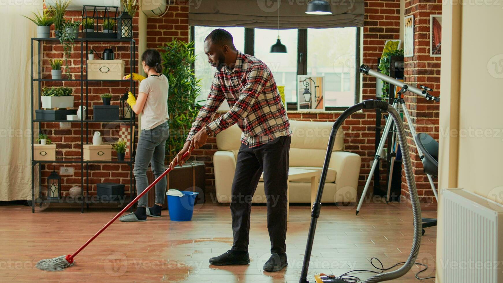 Interracial couple vacuuming floors and cleaning furniture on shelves, sweeping dust or dirt with tools. Life partners using vacuum cleaner with hose, gloves and rags to clean household. House chores. photo