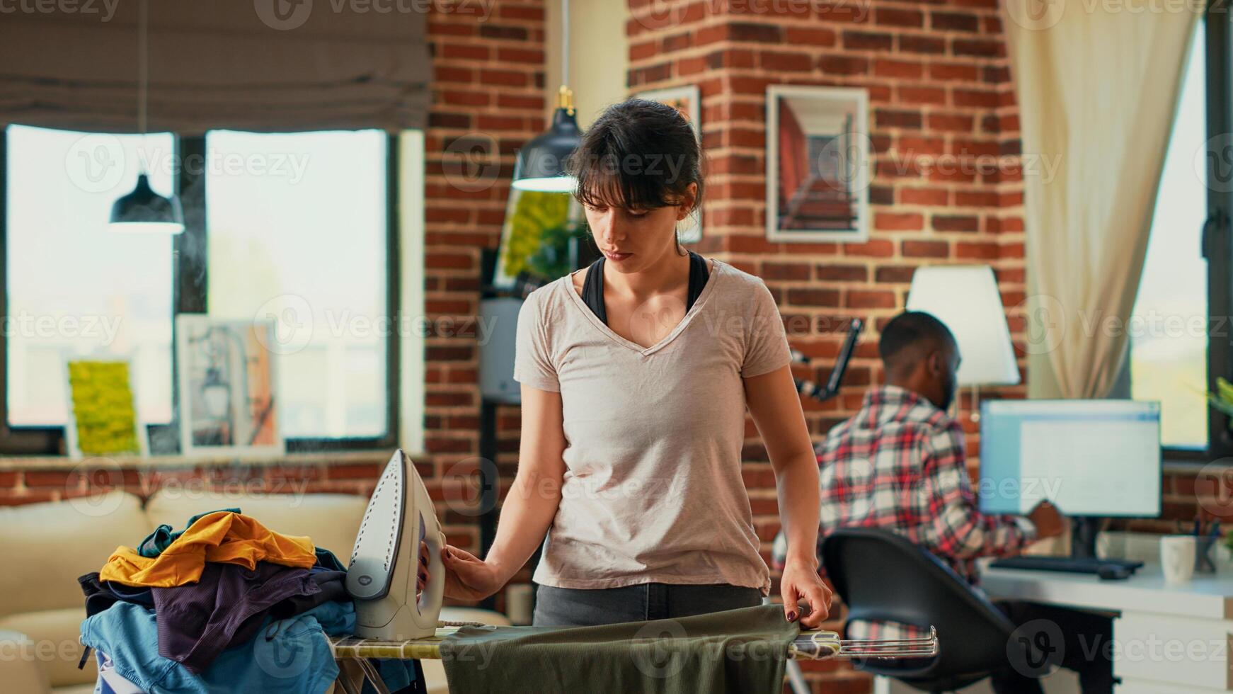 Modern girlfriend using iron with steam to smooth out creases on clothes, ironing pile of laundered garments on ironing board. Casual woman doing spring cleaning at home, housekeeping work. photo