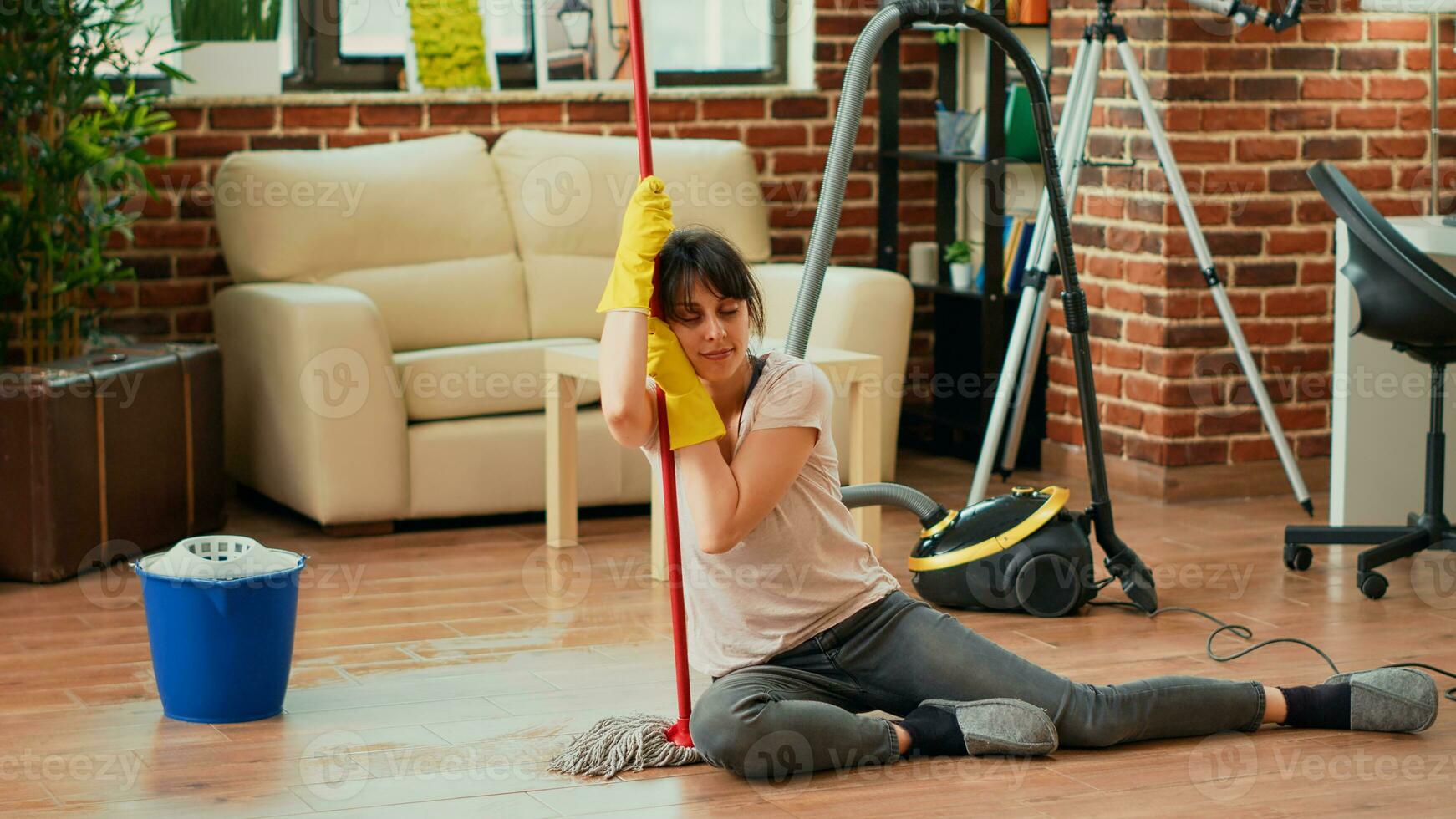 Exhausted woman sitting on living room floors with mop and bucket, feeling satisfied about finishing spring cleaning session. Young person being tired after tidying up household, doing chores. photo