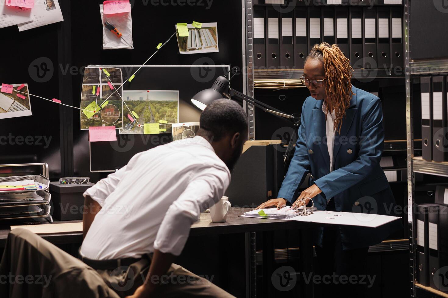 Police office secretary reading crime case file for detective. African american man investigator and woman assistant studying forensic expertise report and working at night time photo