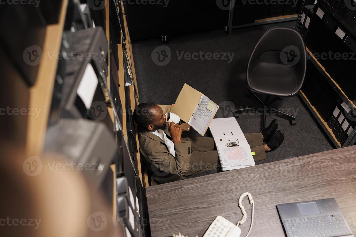 African american cop drinking coffee and analyzing police records on floor. Law enforcement professional working overtime on complex crime case and reading csi expertise reports top view photo