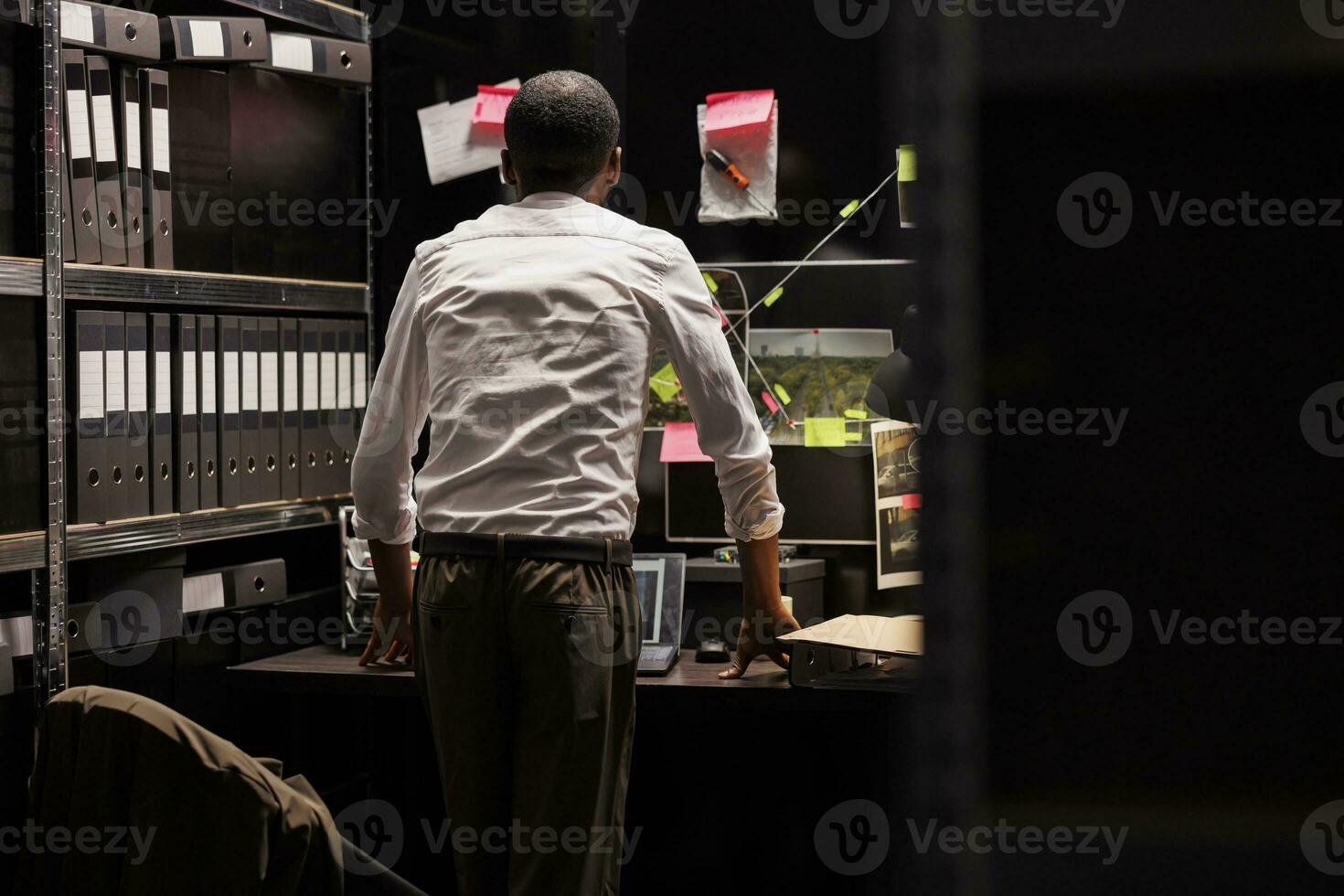 Detective analyzing crime weapon and photos attached to wall, searching insight. African american police investigator looking at evidence and photographs connection map at board