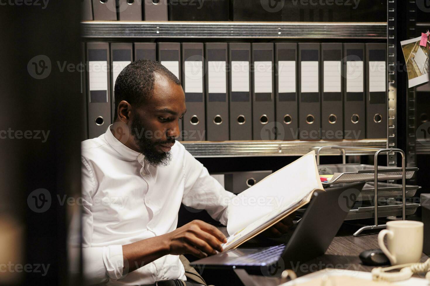 African american prosecutor reading file, preparing for case in police office with archival records. Law enforcement professional analyzing forensic expertise folder at night time photo