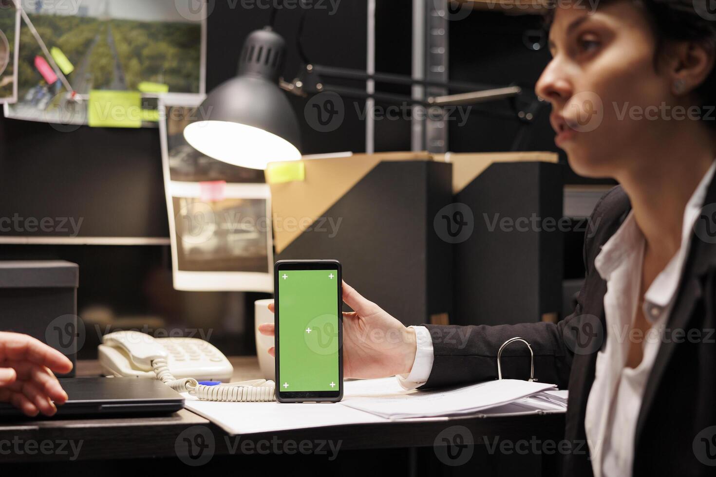 Private detective holding smartphone with green screen mock up display, discussing criminology report with police officer. Investigators working overtime in evidence room. Phone with isolated template photo