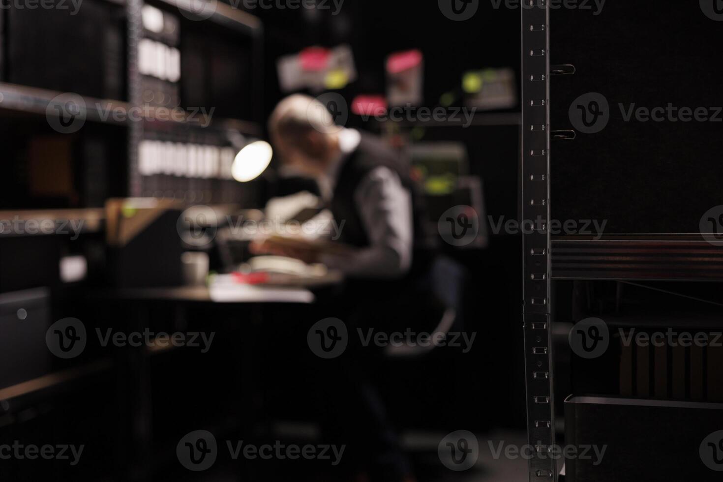 Selective focus of dark room equipped with metallic shelves full with confidential documents. In background old private detective working overhours at criminal case, analyzing investigation evidence photo