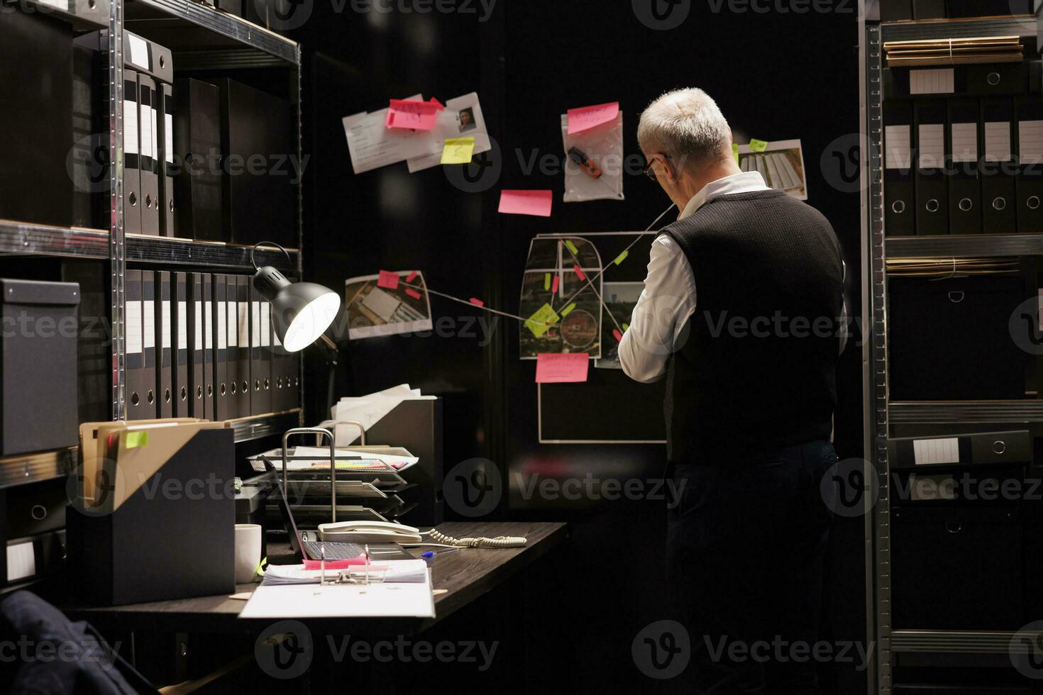 Senior police inspector looking at evidence board, analyzing victim report while working late at night at criminal case in arhive room. Elderly private detective checking criminology files photo