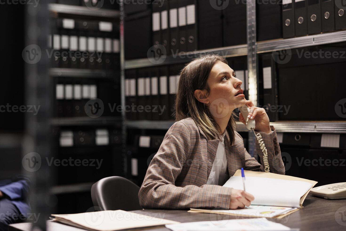 Manager talking at landline phone with remote bookkeeper, discussing accountancy report in storage room. Businesswoman working late at night at administrative catalog in corporate depository photo