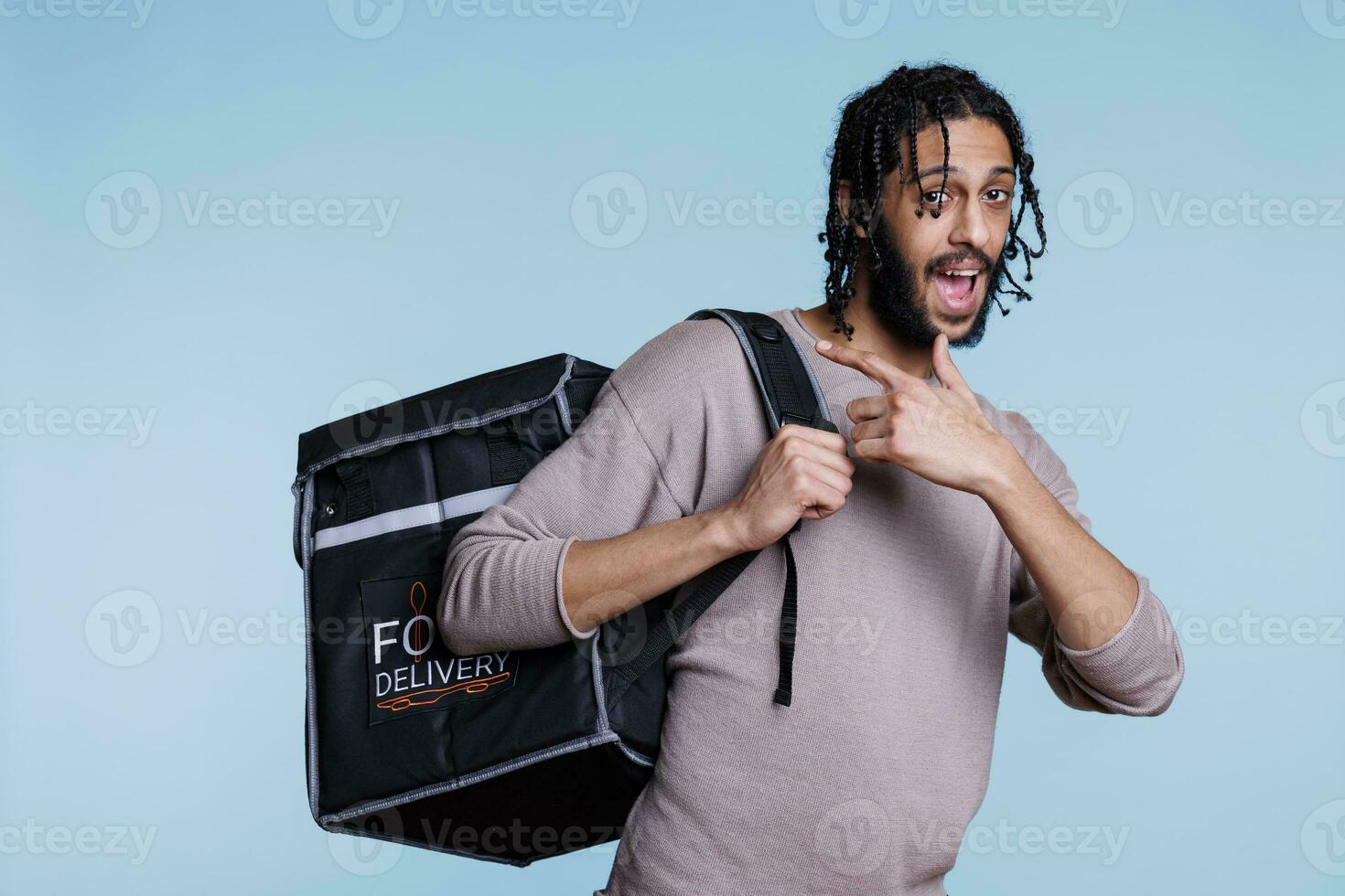 Happy arab restaurant courier pointing at thermal backpack with take away lunch and looking at camera. Young deliveryman with cheerful facial expression carry bag with food portrait photo