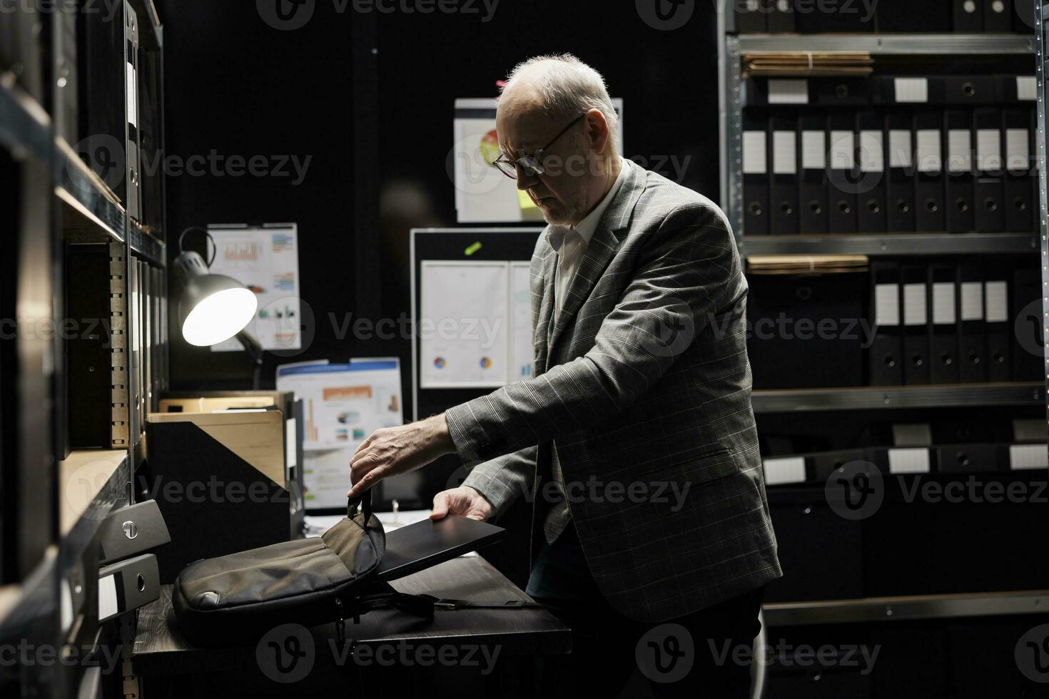 Bureaucratic businessman manager packing laptop up in suitcase bag preparing to clock out. Old bookkeeping executive in accountancy company file cabinet repository office photo