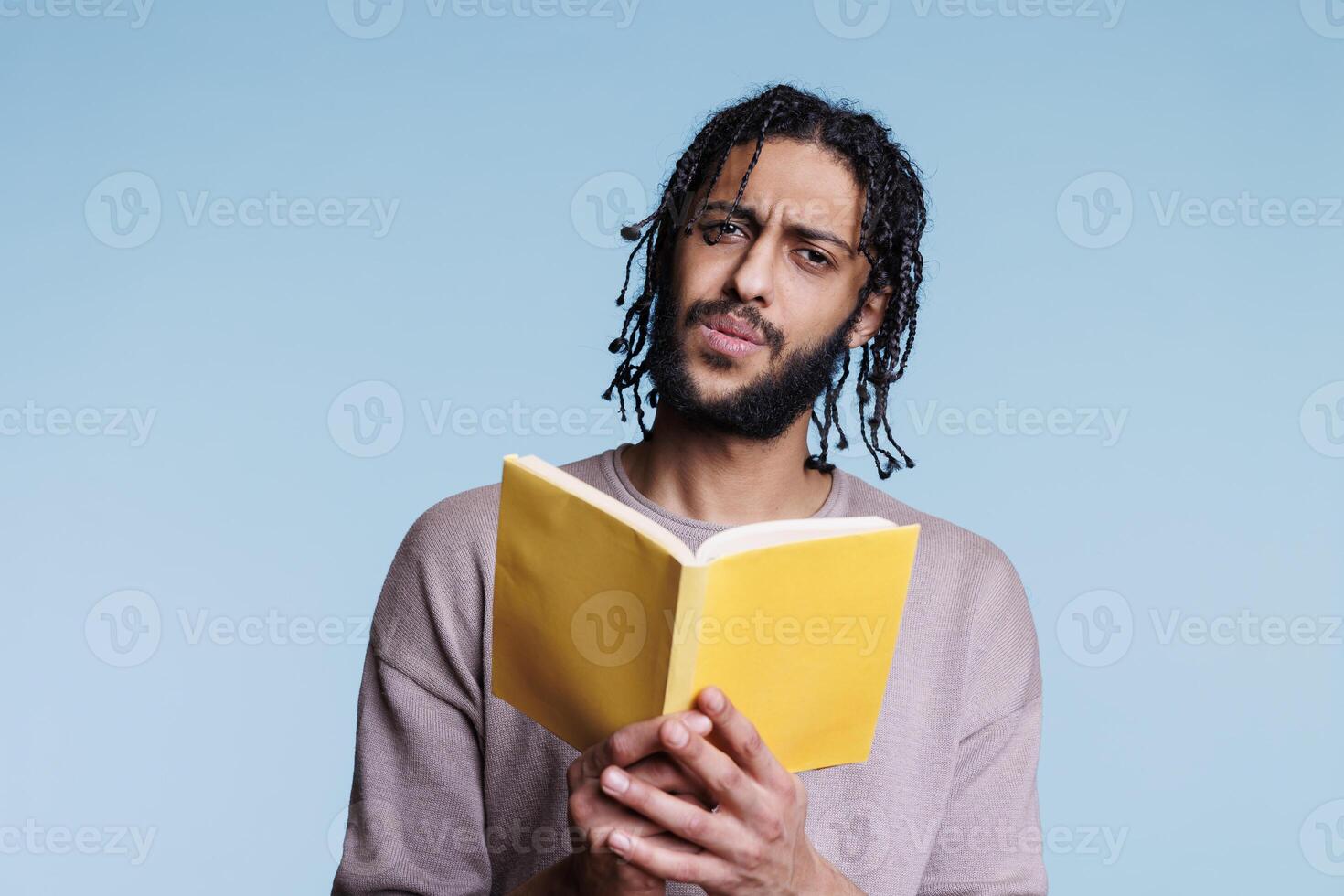 confuso árabe hombre leyendo libro de bolsillo libro y mirando a cámara con perplejo facial expresión. joven cuestionado persona participación abierto tapa blanda libro de texto y aprendizaje retrato foto