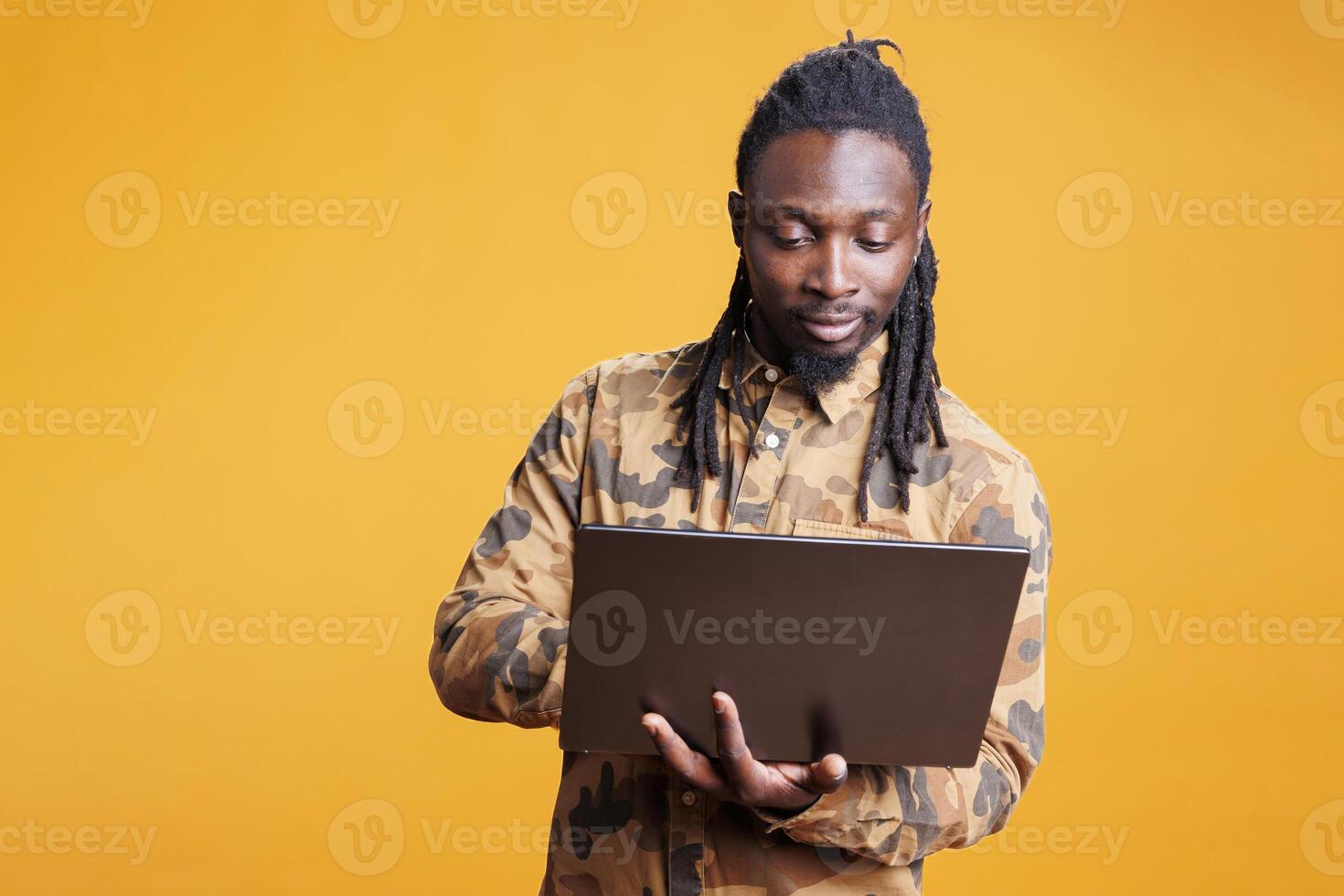 African american man holding laptop computer browsing webpages on internet, standing in studio over yellow background. Young adult using portable device to navigate on social media photo