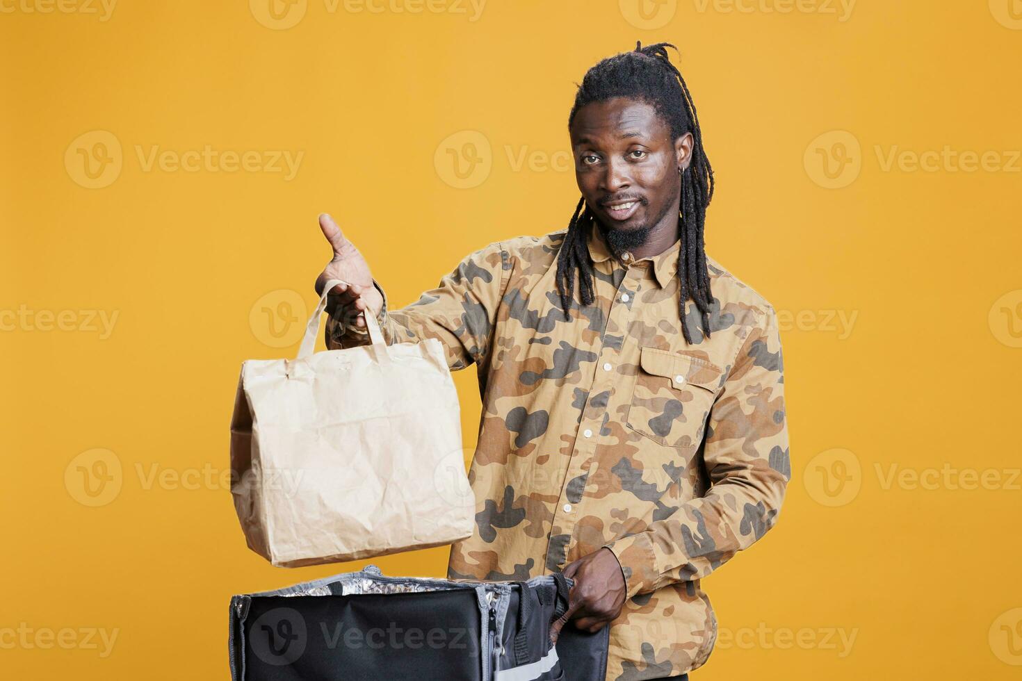 Portrait of pizzeria courier holding paper bag delivering takeaway food to customers during lunch time. African american deliveryman carrying thermal backpack full with fast food in studio photo
