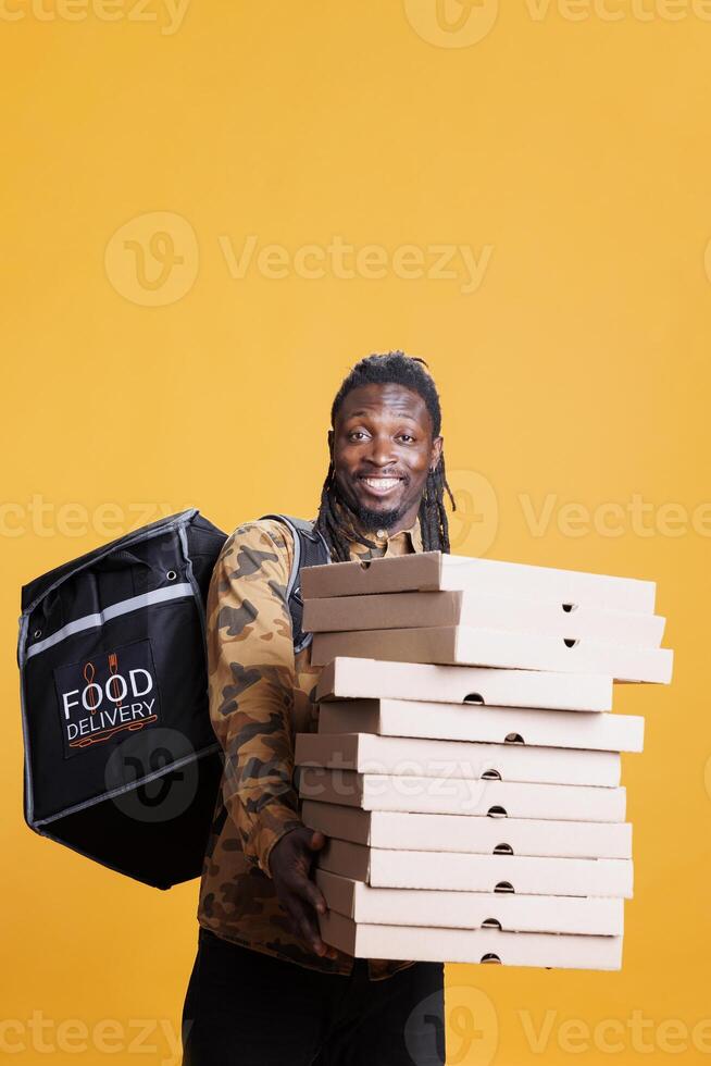 Cheerful deliveryman carrying pizza cardboxes during takeaway delivering, standing in studio over yellow background. Smiling restaurant worker holding thermal backpack. Food service photo