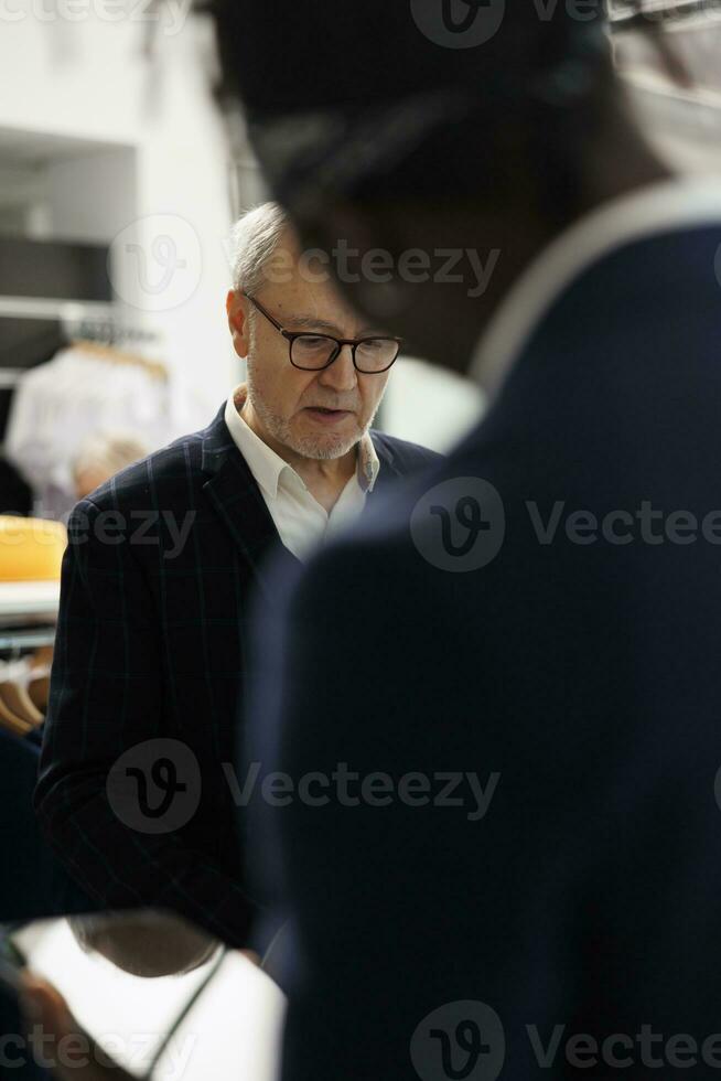 Senior client asking manager to check shirt inventory on tablet computer, wanting to buy formal wear in clothing store. Diverse people discussing merchandise fabric in modern boutique photo