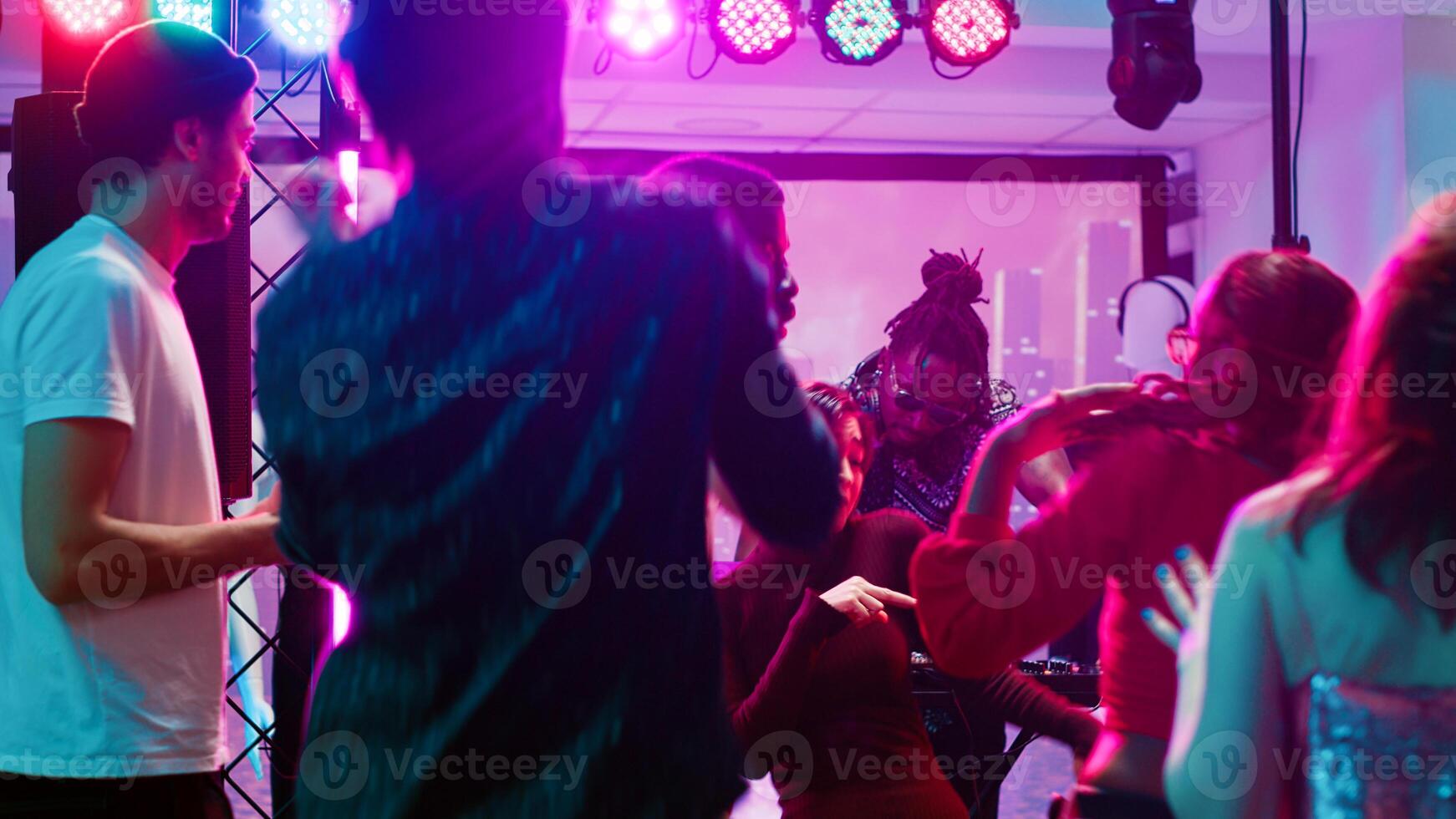 Young people enjoying music at club, dancing on electronic music from DJ mixing station on stage. Funky modern friends clubbing and havng fun with audio sounds, bright spotlights. Tripod shot. photo
