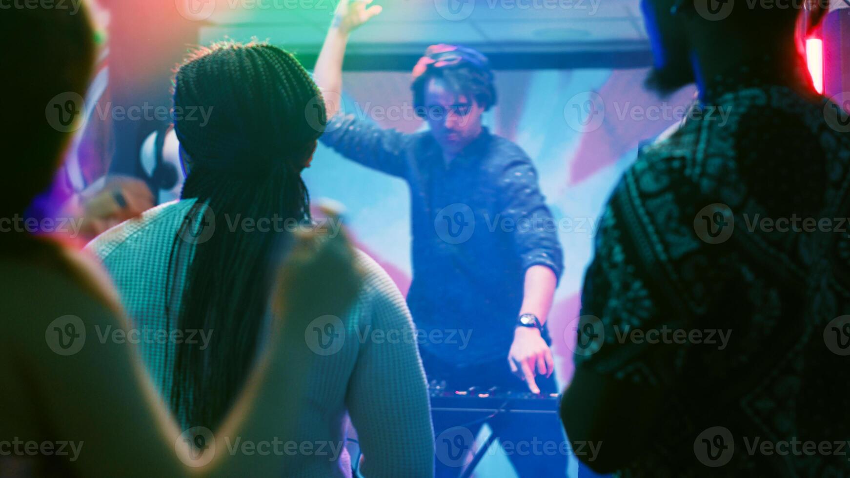 Group of friends dancing in the club, enjoying live performance show with DJ mixing station on nightclub stage. Young people partying together on dance floor with disco lights. Tripod shot. photo