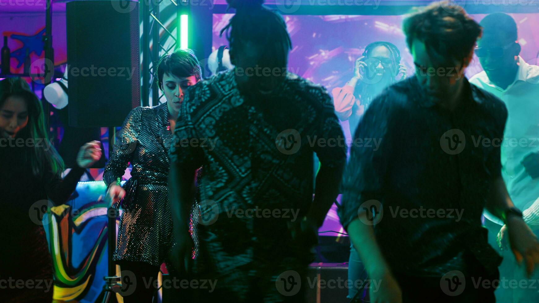 African american DJ mixing music on station, wearing heeadset and having fun with electronic sounds on audio equipment at nightclub. Young woman enjoying clubbing with group of people. photo