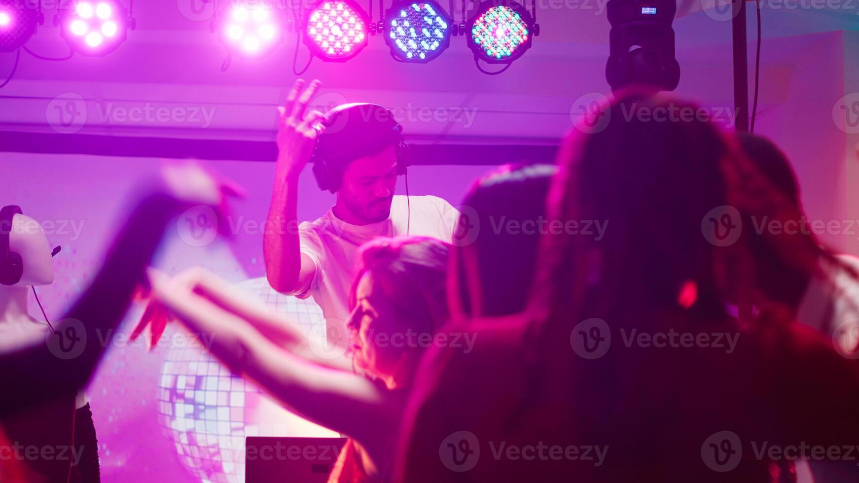 Male DJ partying with people at club, mixing music on audio station to create funky party atmosphere in discotheque. Young man dancing and jumping with friends, having fun on dance floor. photo