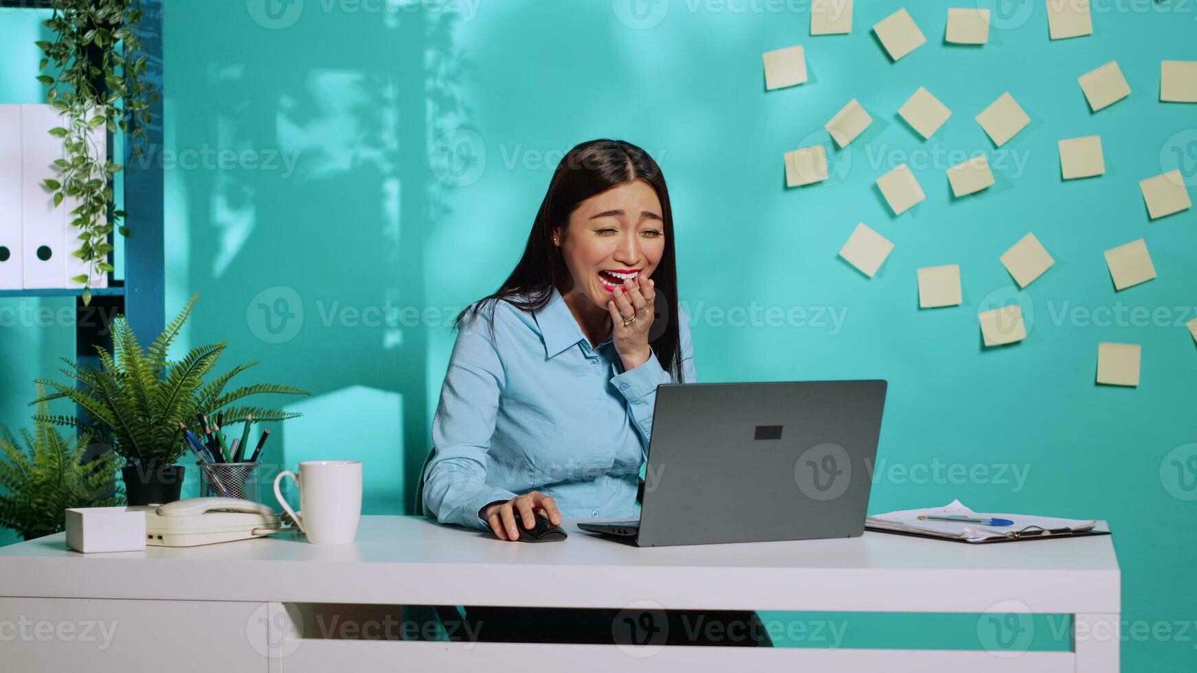 Happy cheerful employee burst out laughing watching laptop screen at work. Lively chipper office clerk having a laugh enjoying herself in colourful relaxed workplace over blue studio background photo
