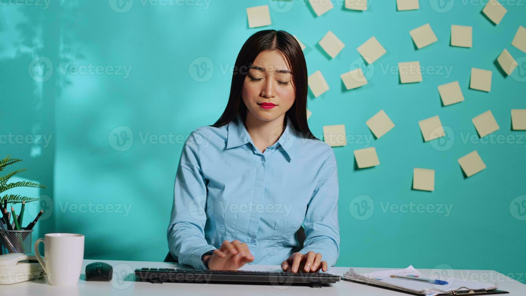 enfocado asiático oficina empleado mecanografía en teclado mientras recepción información desde administración durante en línea videollamada reunión. empleado a vistoso moderno lugar de trabajo escritorio terminado azul estudio antecedentes foto