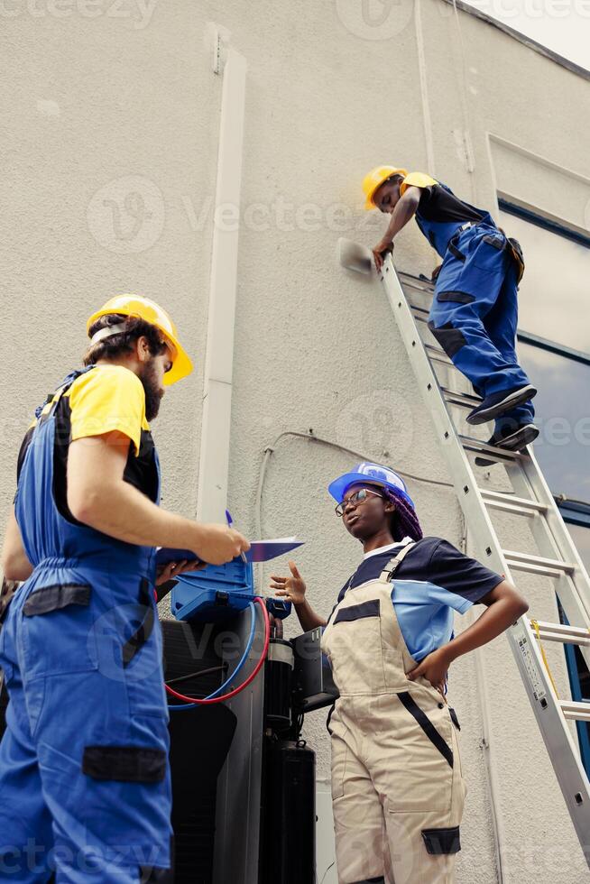 Competent wiremen diverse coworkers commissioned for external air conditioner routine check up, writing findings on clipboard. Professionals doing hvac system investigation of damaged parts photo