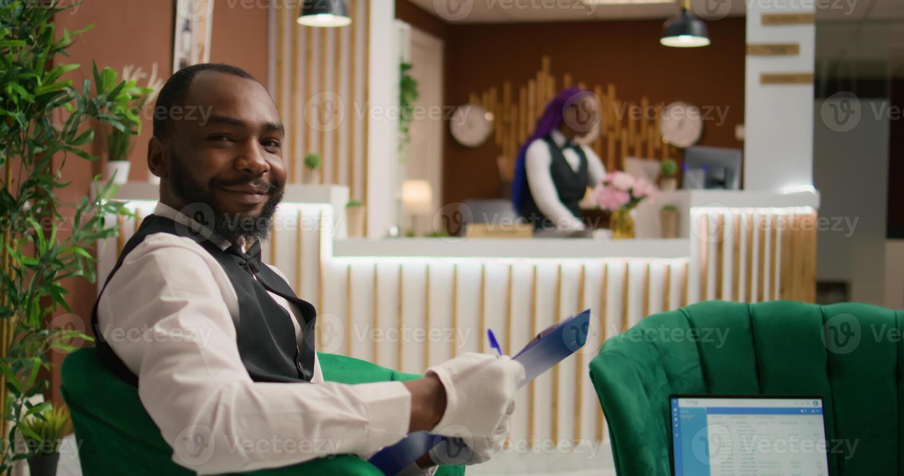 Friendly hotel bellhop reads guests information on clipboard, handing room reservations and providing concierge services in lounge area. African american man writing list of customers. photo