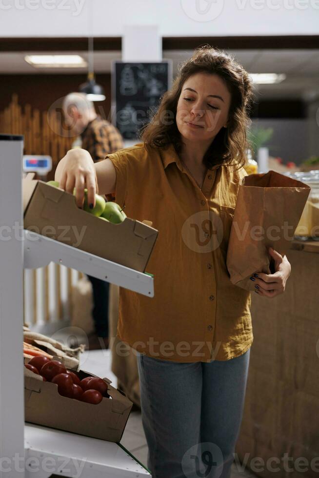 Woman in zero waste shop purchasing farm grown vegetables, picking ripe green apples. Client in plastic free local grocery shop using decomposable paper bag to save planet photo