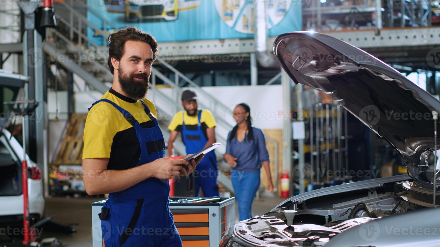 Portrait of smiling skillful mechanic in auto repair shop doing car annual checkup using tablet, looking for damages. Cheerful repairman at work checking to see if vehicle components need replacement photo
