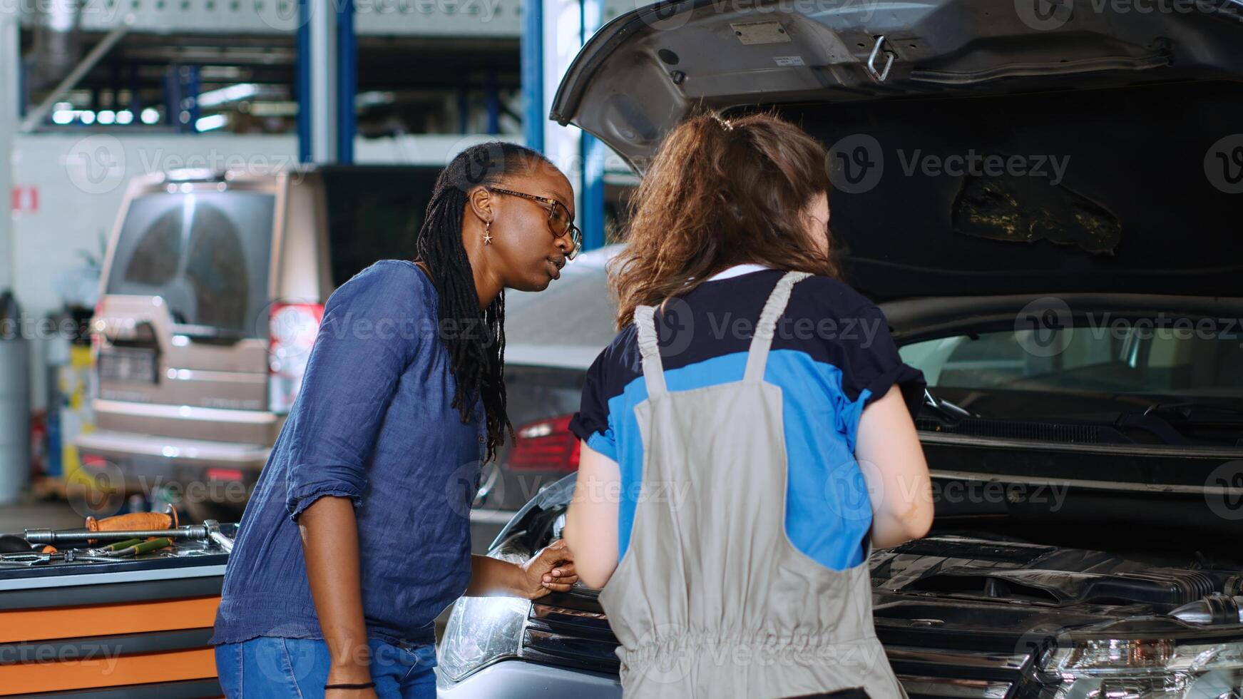 Engineer in garage cleaning opened up vehicle after replacing brake fluid while customer watches. Car service worker wiping automobile leftover leakage and talking with african american client photo