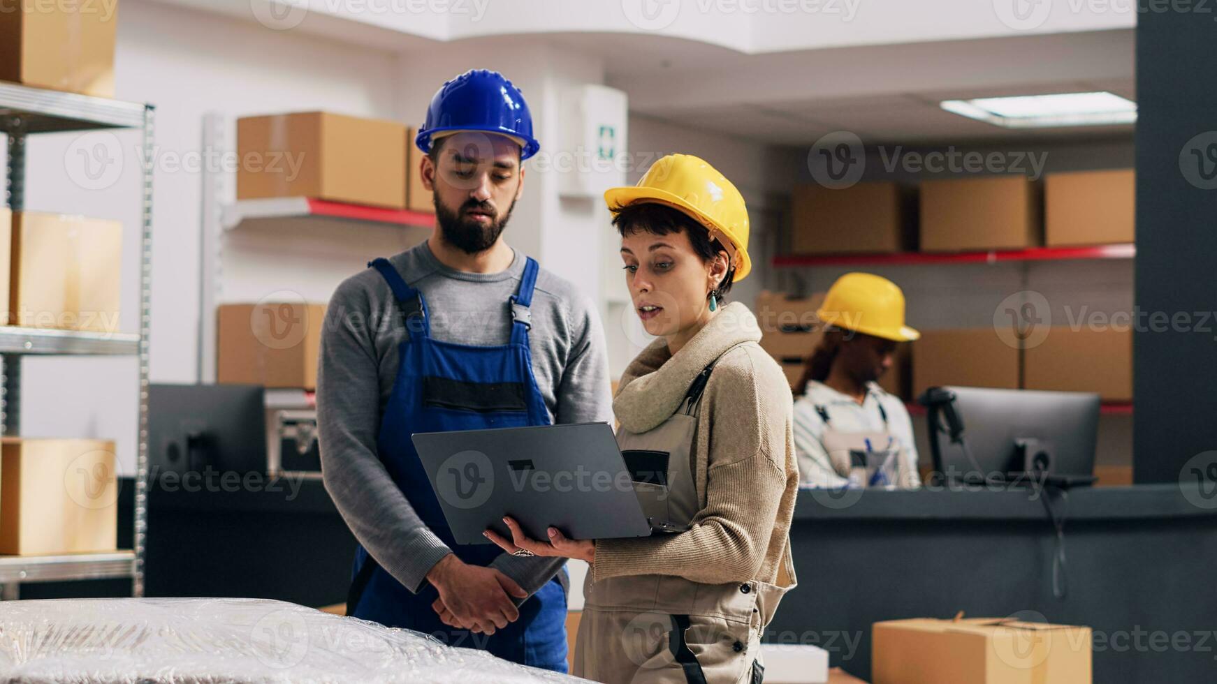 Diverse employees talking about stock logistics in depot, merchandise in boxes placed on storage room racks. Man and woman wearing overalls and hardhats using laptop in warehouse space. photo