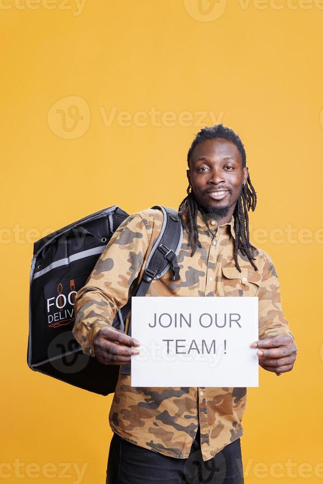 Pizzeria worker holding job offer sign, recruiting people for food delivery in studio over yellow background. African american man asking people to attend courier interview. Takeout concept photo