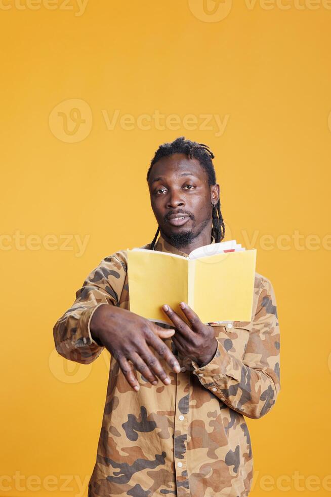 Portrait of serious reader holding literature book, reading fiction story in studio over yellow background. Concentrated african american man learning before start working at university homework photo