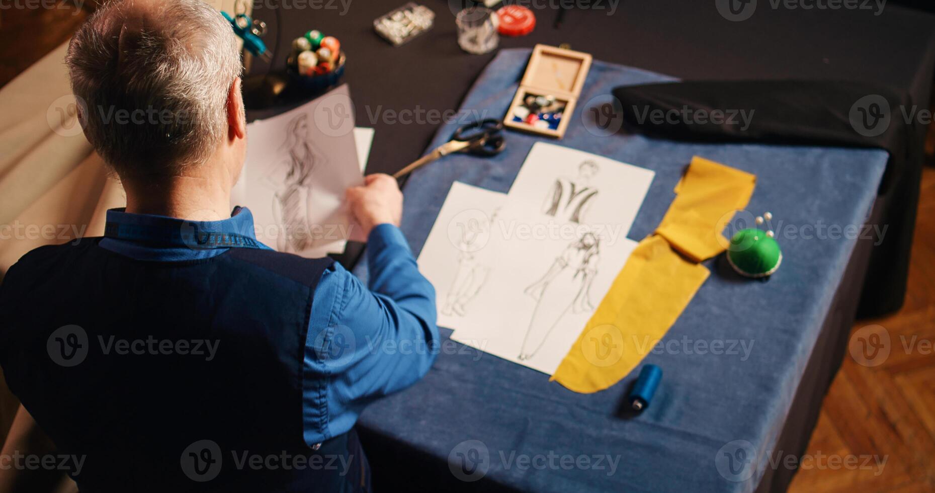 Old designer choosing material comparing sketches, using luxury clothing items for fashion collection. Senior tailor using modern sewing equipment in handcraft atelier. Handheld shot. photo