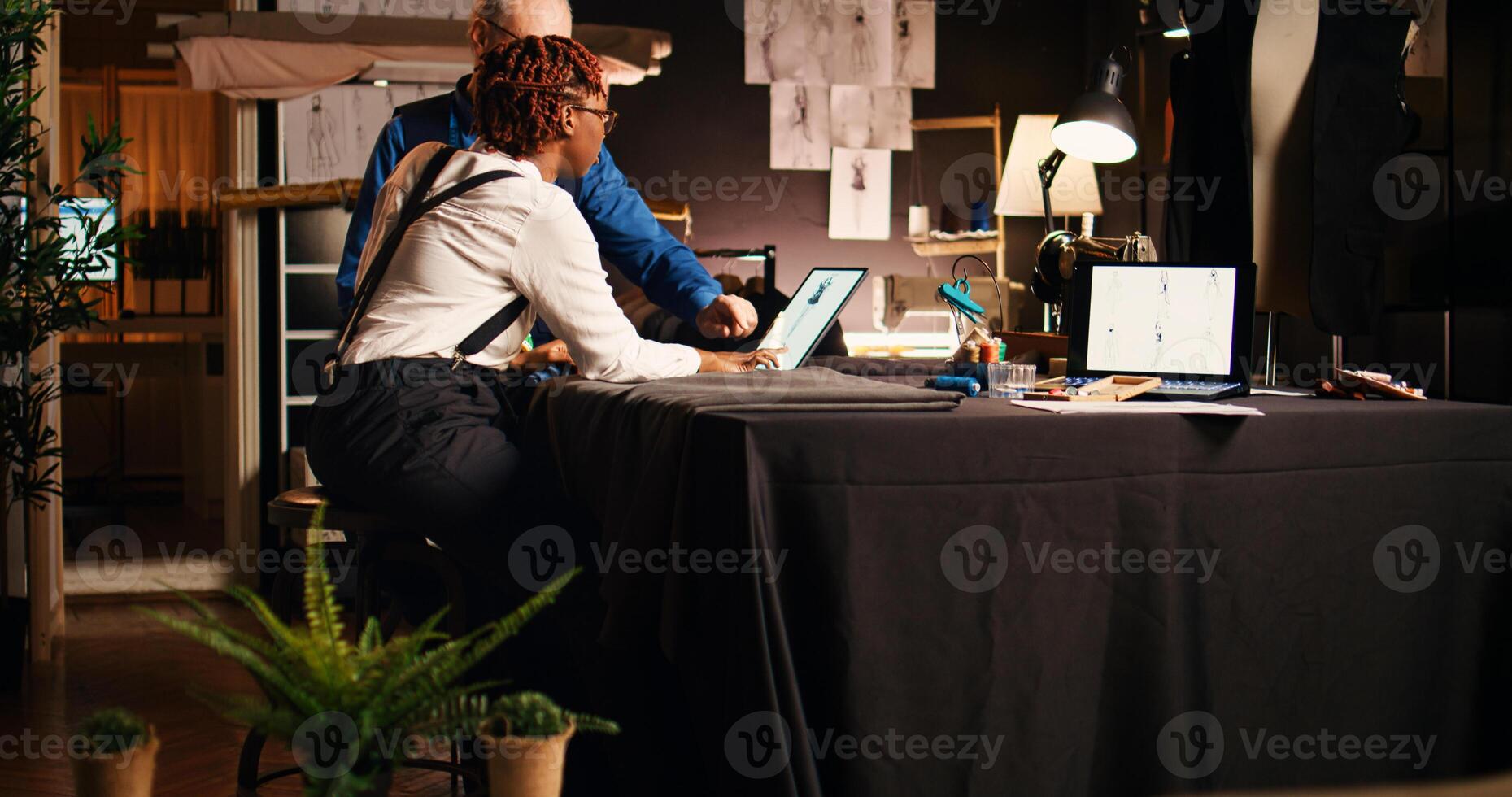 Senior tailor and apprentice in atelier discussing about new clothing models with sketches, handmade concept. Experienced craftsman teaching trainee to use sewing machine, needlework. photo