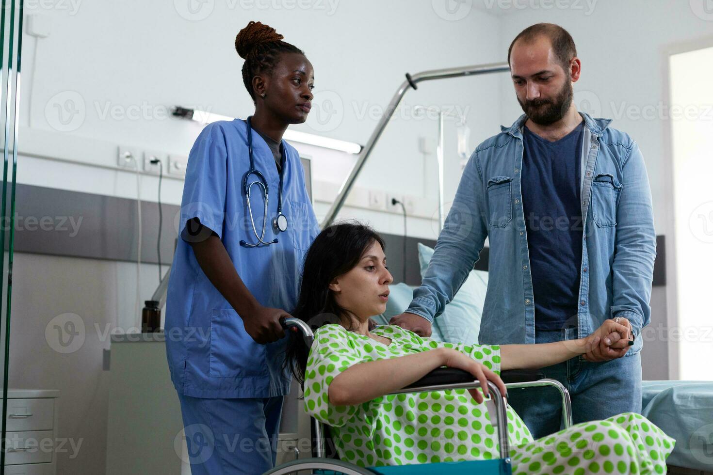 African american nurse taking pregnant woman in wheelchair to maternity room to delivery baby. Patient with pregnancy preparing for childbirth while being comforting by husband in hospital ward photo