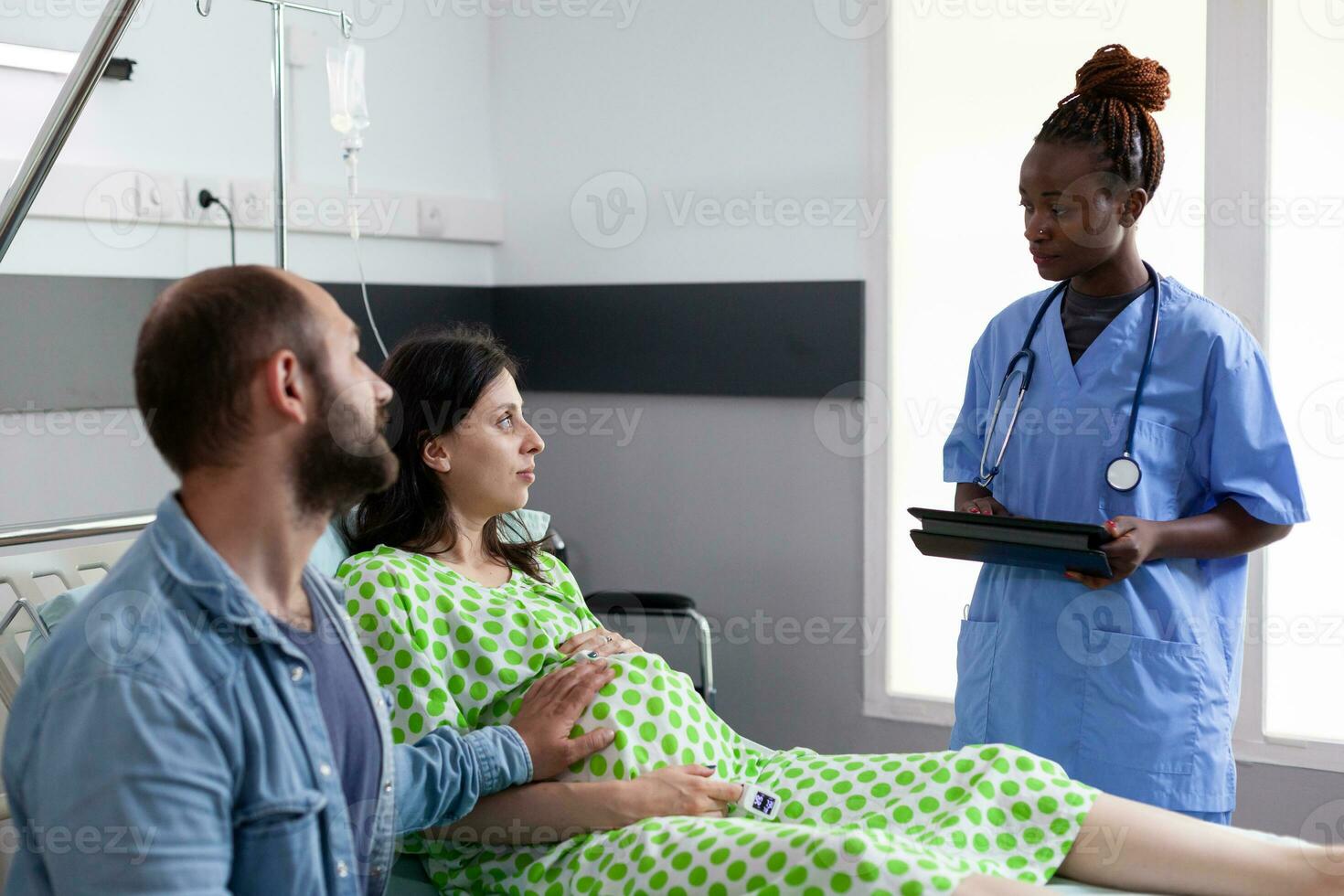 African american nurse explaining labor process to pregnant woman, preparing couple for child delivery in hospital ward. Patient with pregnancy lying in bed while husband comforting about motherhood photo