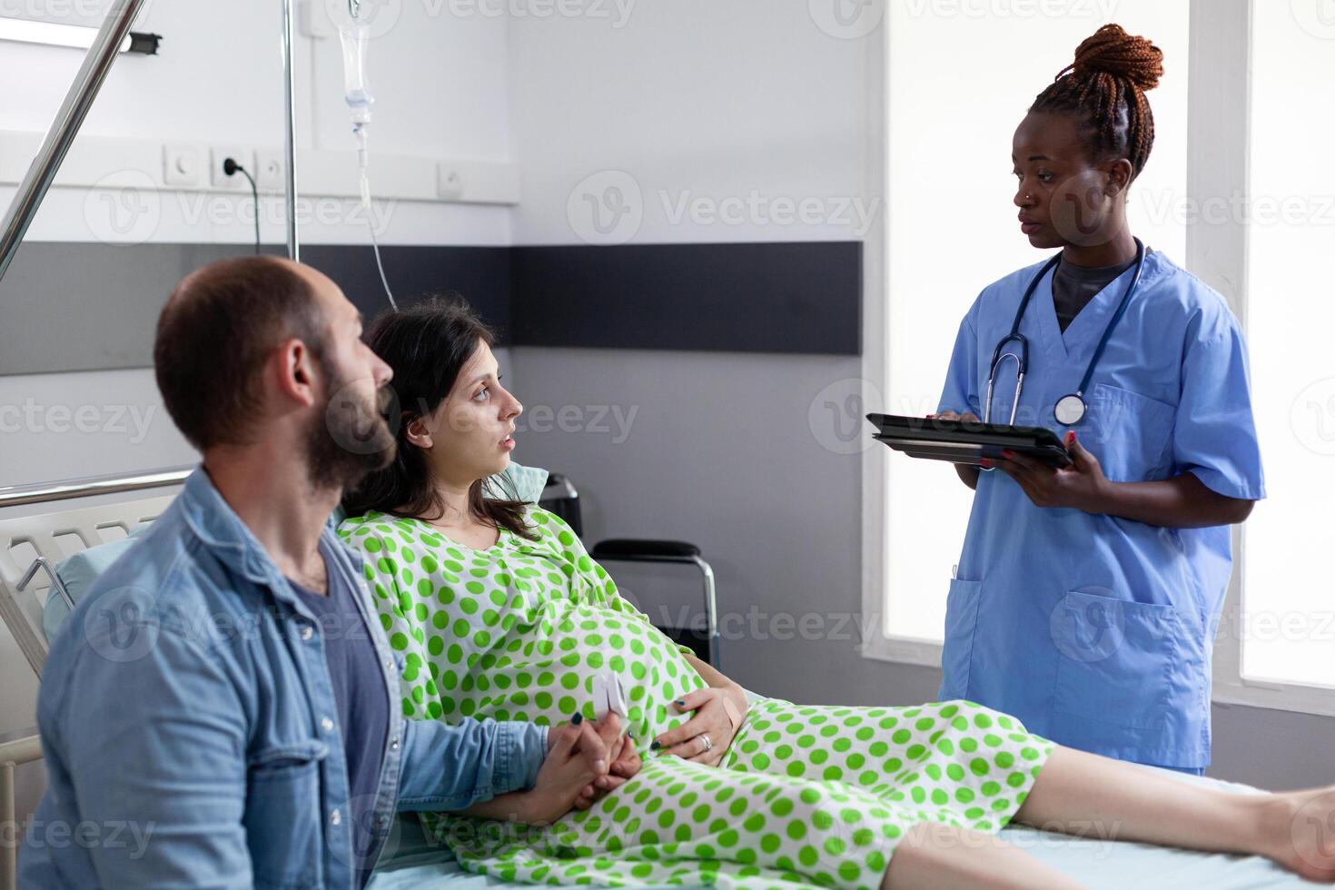 Future parents discussing with african american nurse before childbirh in hospital ward. Medical assistant typing patient contractions level on tablet computer, explaining pregnancy labor process photo