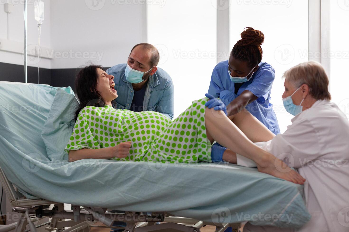 Diverse nurse and medic helping with childbirth in hospital ward. Mother lying in bed having painful contractions giving birth to baby while father standing beside her comforting in maternity clinic photo
