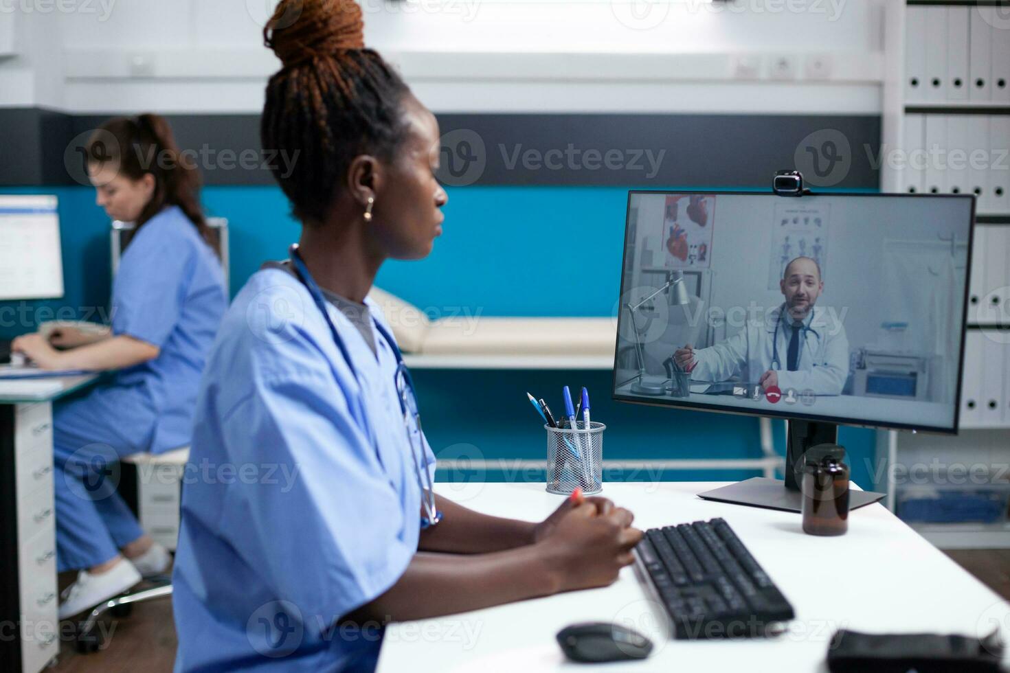 Medical team on internet videoconference telehealth video call in busy clinical office. African american nurse at hospital desk consulting with general practitioner about patient case photo