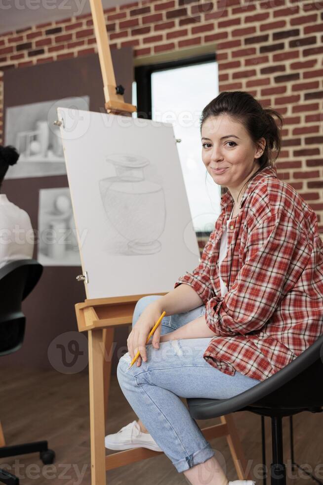 Happy young woman smiling at camera while sitting at easel during fun and creative art class, learning basics of drawing, trying something new. Student girl enjoying arts activities photo