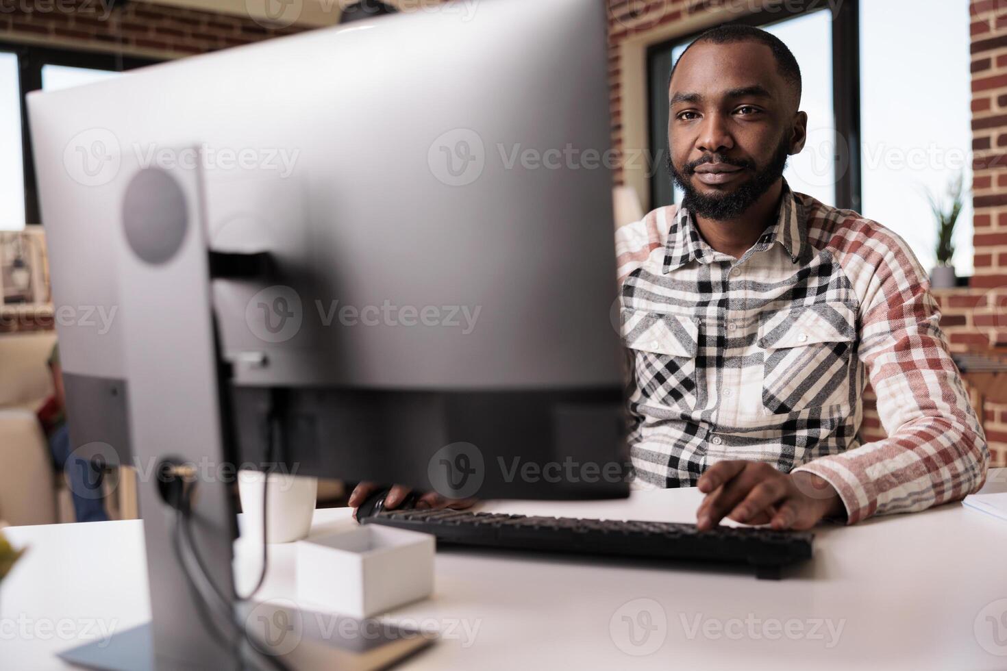 Confident african american programmer looking at computer screen and typing while working remote from home living room. Selective focus on freelancer using pc for remote job sitting at desk. photo