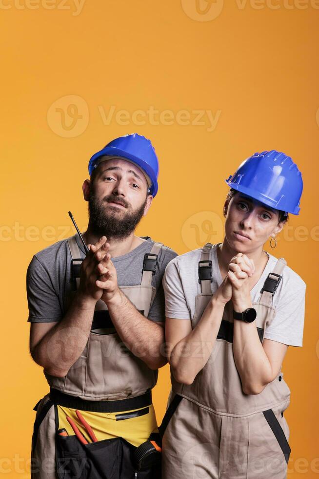 Religious repairman and woman praying to god for good projects, begging for help. Team of engineering experts holding prayer hands to ask for fortune and luck, wearing overalls and helmets. photo