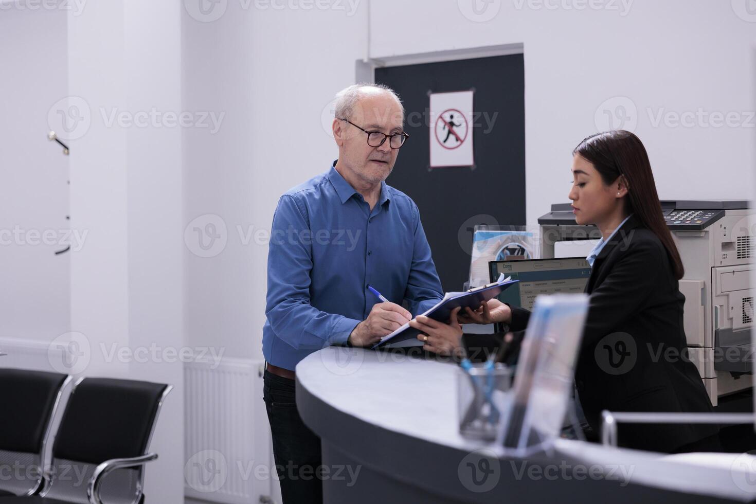 Elderly patient discussing disease expertise with asian receptionist while signing medical paper, standing at hospital counter in hospital waiting area. Health care service and concept photo