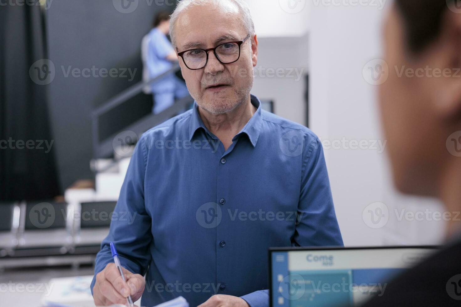 Old patient discussing illness expertise with asian reception worker while signing medical documents, standing at hospital counter in hospital waiting room. Health care service and concept photo