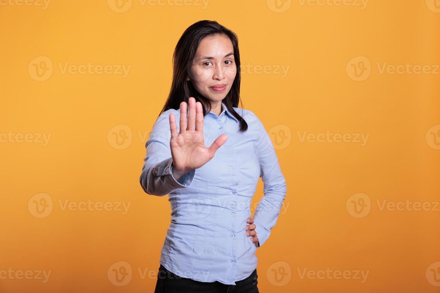 Negative asian woman doing refusal gesture with hand in front of camera, posing in studio over yellow background. Serious young adult showing stop gesture, being in denial and expressing rejection. photo