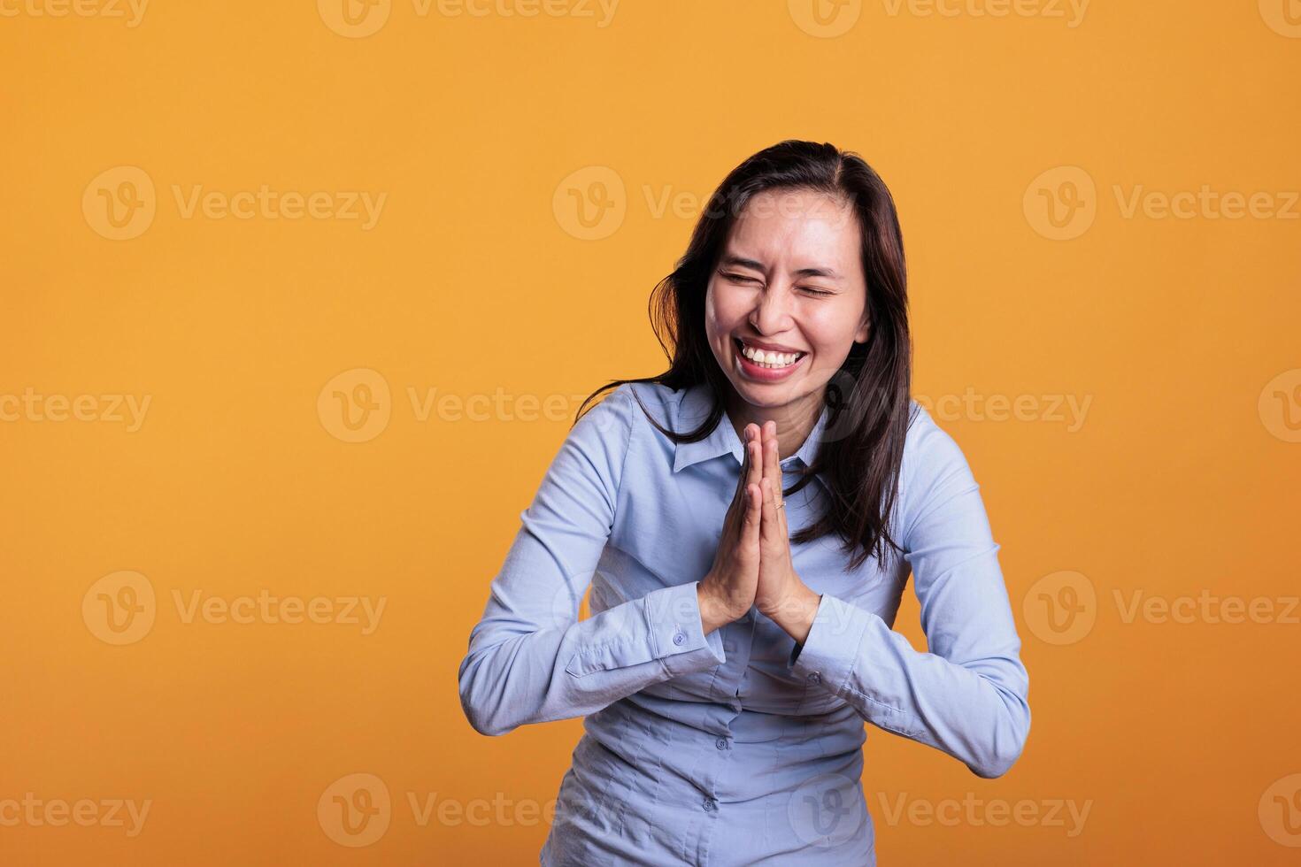 Cheerful filipino woman praying to god asking for forgiveness, posing with palms together in studio over yellow background. Positive hopeful young adult showing religious spiritual gesture photo